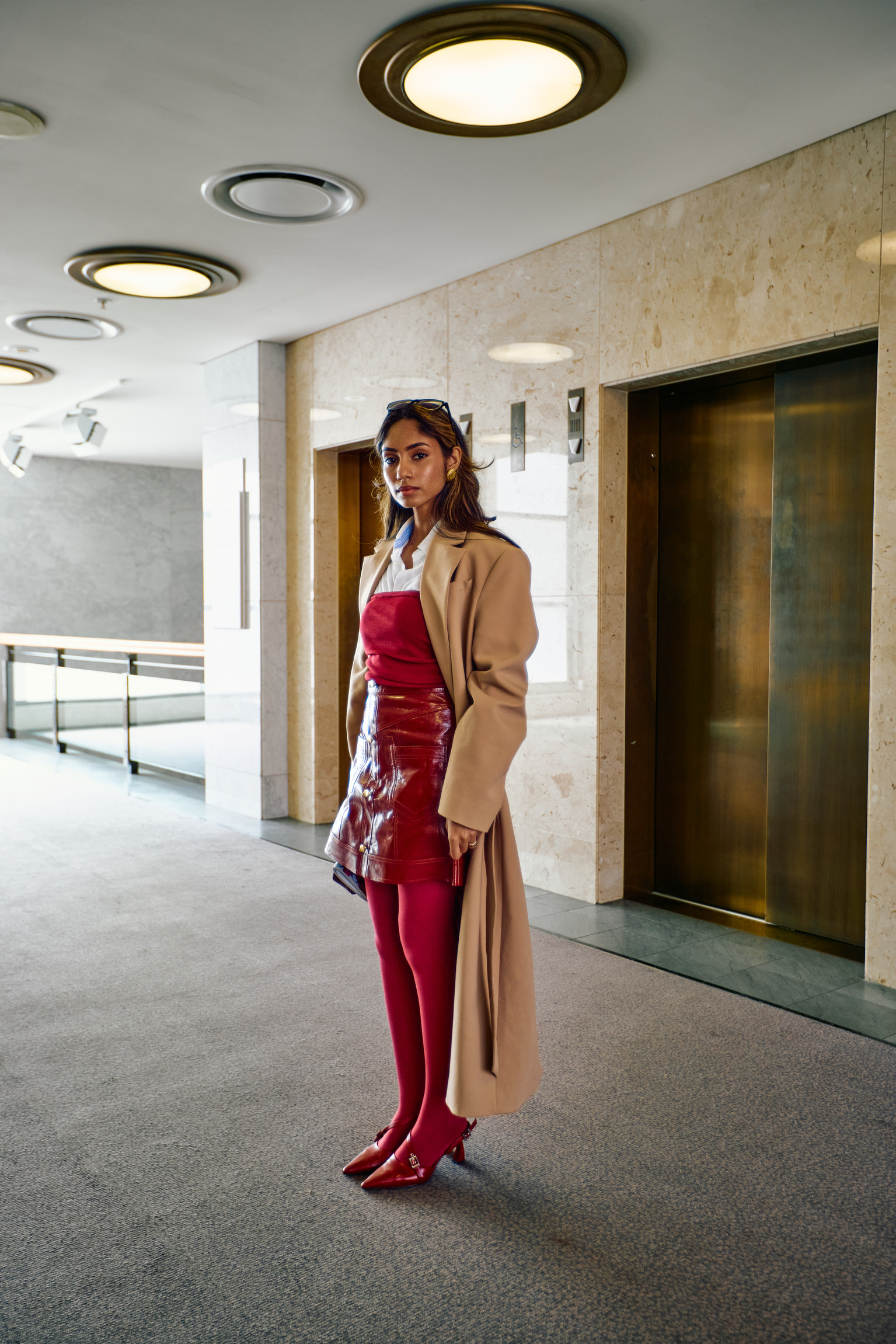 Stylish woman in red dress and beige coat stands in modern building hallway with elevators and bright lighting for fashion editorial.